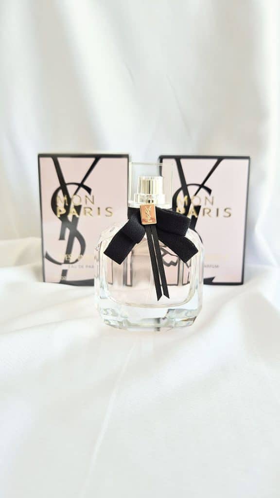 Vue de face du flacon de l'eau de parfum Mon Paris d'Yves Saint Laurent, orné de son nœud lavallière noir et du logo doré YSL, posé sur un drapé blanc lumineux devant son packaging rose et noir.