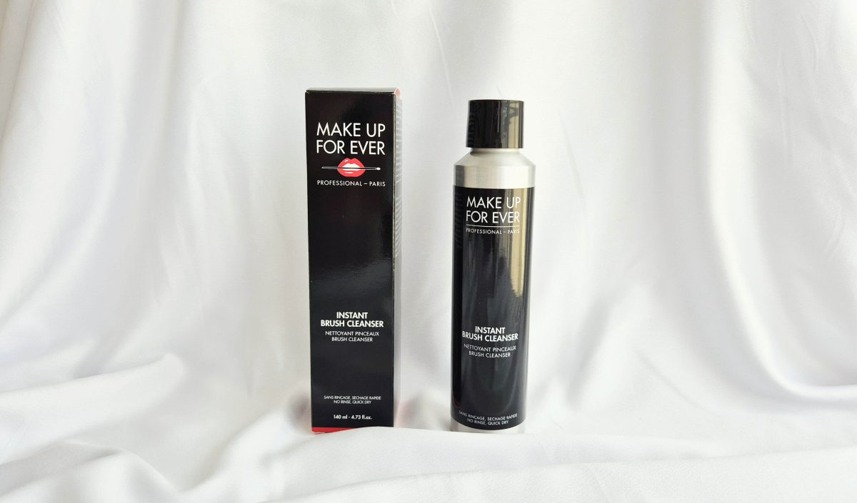 Flacon noir en métal et boîte en carton du nettoyant pour pinceaux Instant Brush Cleanser de Make Up For Ever sur fond blanc en tissu.