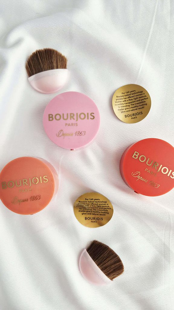 Vue de dessus de trois boîtiers fermés de blush Boîte Ronde Bourjois, accompagnés de deux petits pinceaux applicateurs incurvés et de deux opercules dorés sur un tissu blanc.