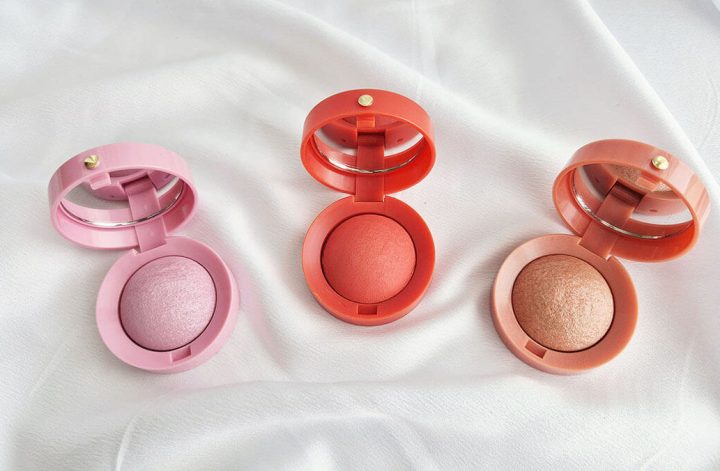 Trois blushs Boîte Ronde de Bourjois ouverts dans les teintes rose, corail et pêche, posés sur un tissu en satin blanc.