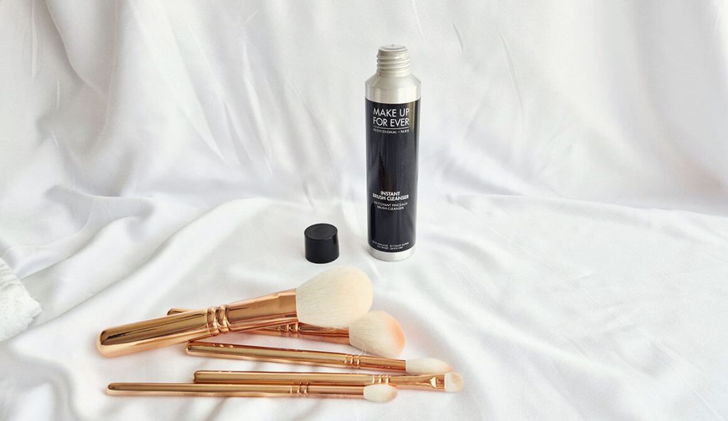 Flacon ouvert d'Instant Brush Cleanser de Make Up For Ever posé derrière plusieurs pinceaux Zoeva Rose Gold sales avec des résidus de maquillage orange sur les poils blancs.