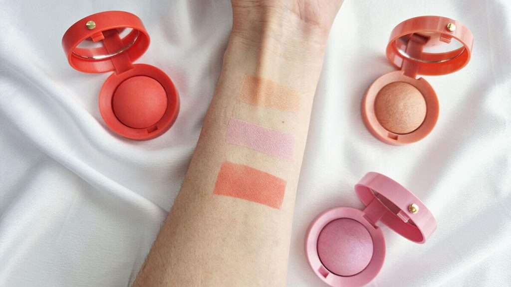 Test de pigmentation avec trois swatches de fards à joues Bourjois sur un avant-bras, entouré des petits boîtiers ronds ouverts.