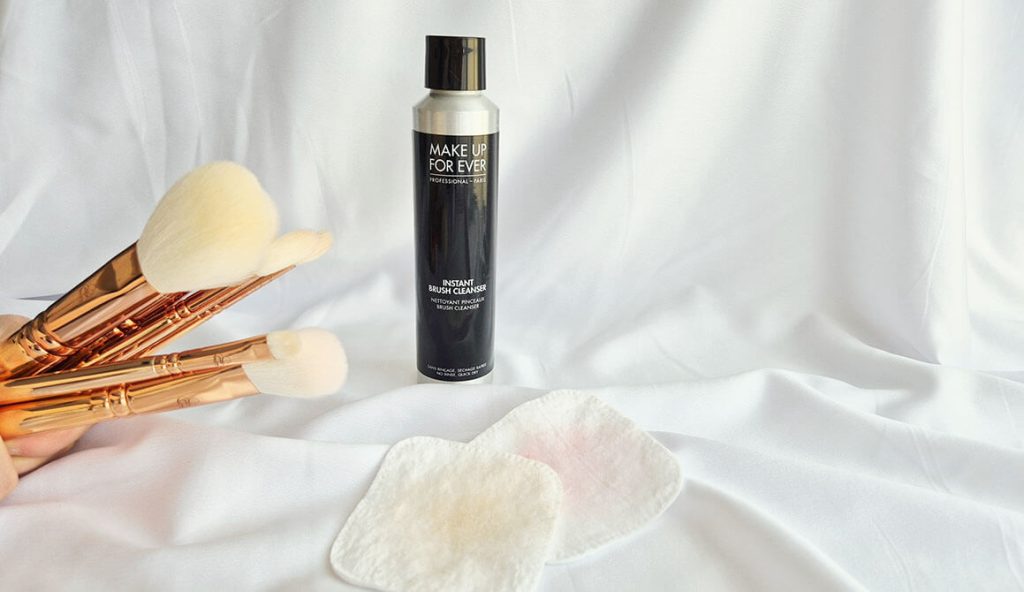 Flacon de lotion sans rinçage posé derrière deux disques de coton usagés contenant des traces de maquillage, à côté de pinceaux Zoeva Rose Gold tenus à la main.