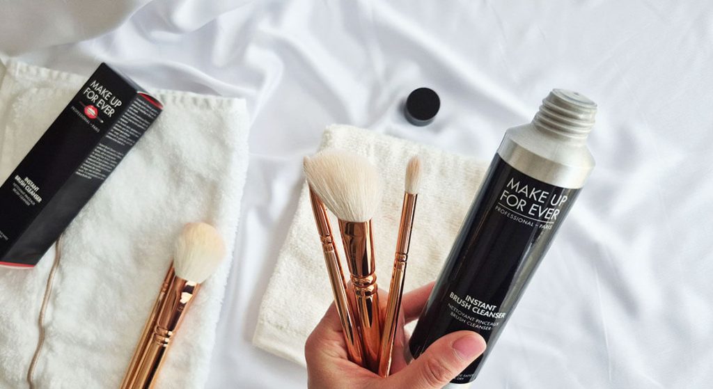 Main tenant le flacon Instant Brush Cleanser de Make Up For Ever et trois pinceaux Zoeva Rose Gold au-dessus de gants de toilette blancs.
