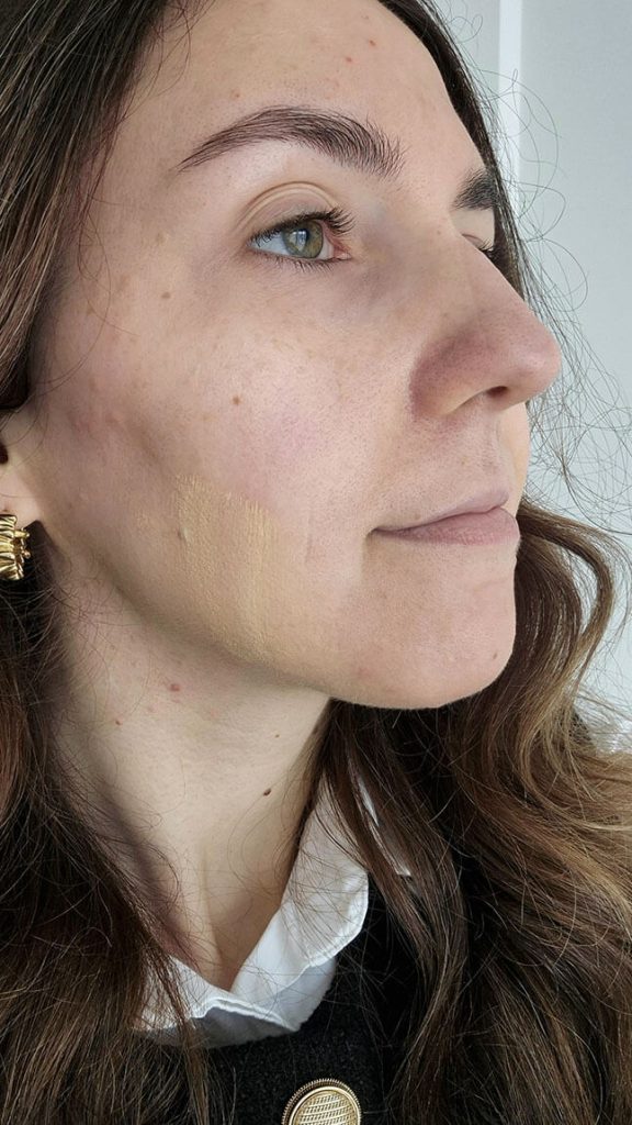 Application d'une touche de fond de teint Haus Labs de couleur claire et jaune sur la joue d'une femme de profil.
