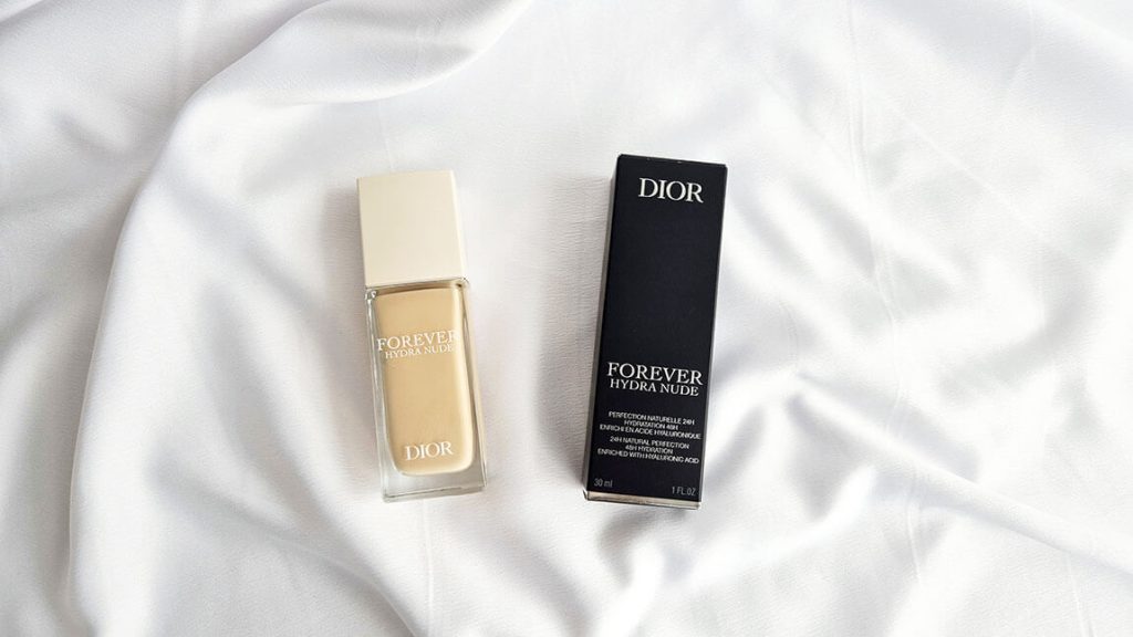 Le flacon de fond de teint Dior Forever Hydra Nude et sa boîte d'emballage en carton noir présentés allongés côte à côte sur un tissu blanc satiné.
