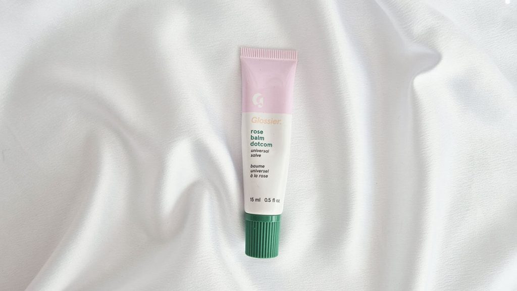 Tube de baume à lèvres Balm Dotcom Glossier teinte Rose avec bouchon vert posé sur un tissu blanc.