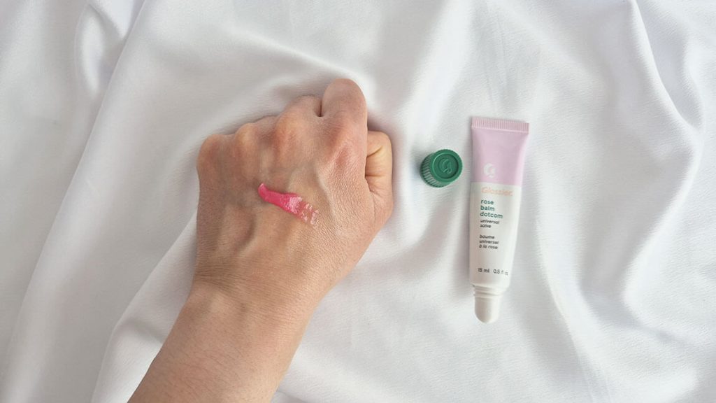 Swatch de la teinte Rose du baume Balm Dotcom Glossier à côté du tube sur fond blanc.